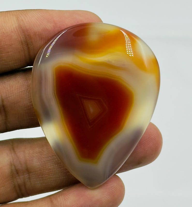 May include: A polished, teardrop-shaped cabochon gemstone featuring a vibrant central core of deep reddish-brown surrounded by concentric bands of translucent amber, golden yellow, and creamy white, with a smooth, glossy, reflective surface.