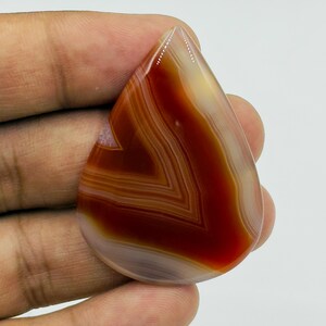 Pode incluir: Uma pedra preciosa de &aacute;gata polida em forma de l&aacute;grima com faixas conc&ecirc;ntricas de castanho-avermelhado profundo, creme transl&uacute;cido e branco. A pedra tem uma superf&iacute;cie lisa e brilhante com padr&otilde;es naturais de faixas vis&iacute;veis em toda a face.