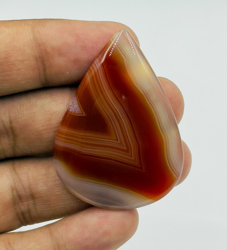 Pode incluir: Uma pedra preciosa de &aacute;gata polida em forma de l&aacute;grima com faixas conc&ecirc;ntricas de castanho-avermelhado profundo, creme transl&uacute;cido e branco. A pedra tem uma superf&iacute;cie lisa e brilhante com padr&otilde;es naturais de faixas vis&iacute;veis em toda a face.