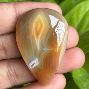 May include: A polished, teardrop-shaped gemstone with a swirling pattern of tan, white, and orange hues. The stone is held in a hand, showcasing its smooth, reflective surface and natural color variations. The gemstone's unique design makes it a beautiful piece.