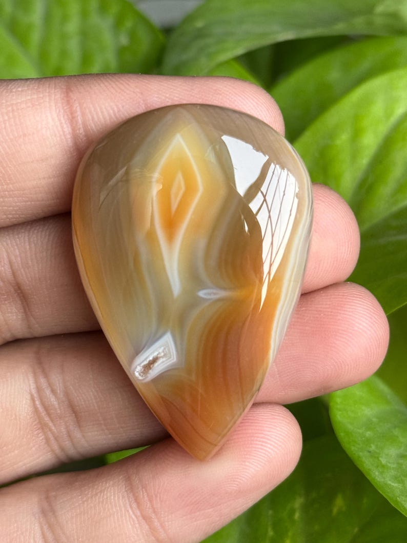 May include: A polished, teardrop-shaped gemstone with a swirling pattern of tan, white, and orange hues. The stone is held in a hand, showcasing its smooth, reflective surface and natural color variations. The gemstone's unique design makes it a beautiful piece.