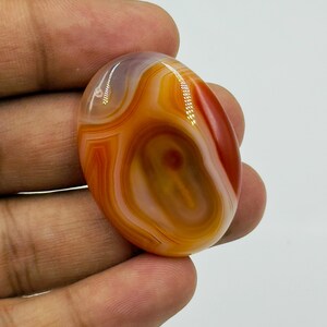 May include: A polished oval-shaped agate gemstone featuring concentric bands of translucent orange, deep red, and creamy white colors. The stone has a smooth, glossy surface with natural layered patterns and a distinct dark red center point.
