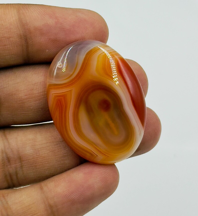 May include: A polished oval-shaped agate gemstone featuring concentric bands of translucent orange, deep red, and creamy white colors. The stone has a smooth, glossy surface with natural layered patterns and a distinct dark red center point.