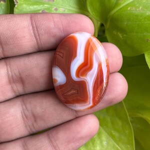 May include: An oval-shaped cabochon gemstone featuring a polished surface with distinct, swirling bands of deep burnt orange, terracotta, and opaque white colors, showcasing a smooth, glossy finish and a symmetrical, rounded edge profile.