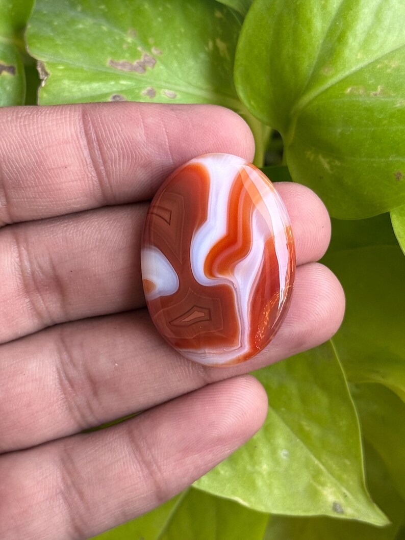 May include: An oval-shaped cabochon gemstone featuring a polished surface with distinct, swirling bands of deep burnt orange, terracotta, and opaque white colors, showcasing a smooth, glossy finish and a symmetrical, rounded edge profile.