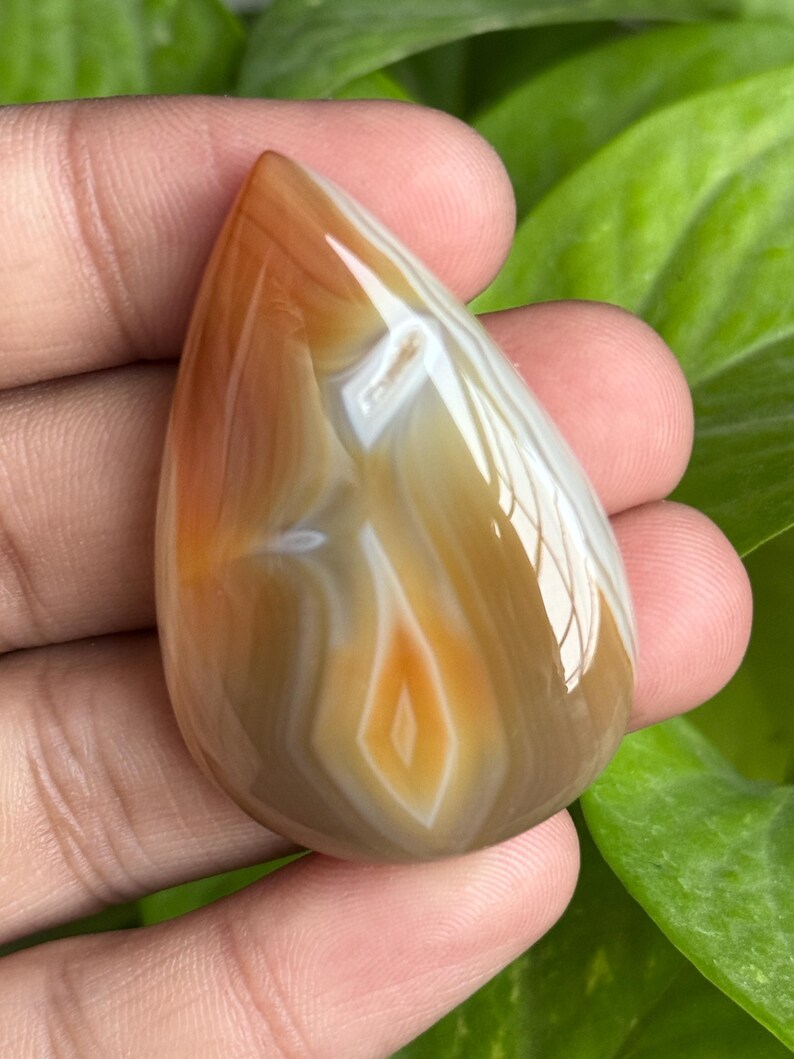 May include: A polished, teardrop-shaped gemstone with a mix of brown, white, and orange hues. The stone has a glossy finish and a layered pattern, with a central diamond-like design. The gemstone is held in a hand against a backdrop of green leaves.