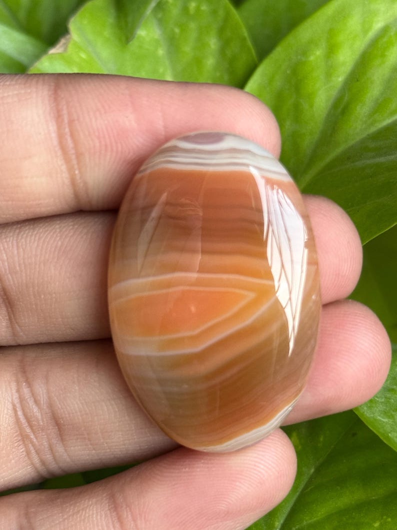 May include: An oval-shaped gemstone with a polished surface. The stone features bands of orange, brown, and white, creating a layered effect. The gemstone is held in a hand, with green foliage in the background.