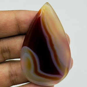 May include: A polished, teardrop-shaped agate gemstone featuring smooth, flowing bands of deep maroon, translucent cream, and soft amber. The stone has a glossy, reflective surface with distinct, organic color transitions throughout its structure.