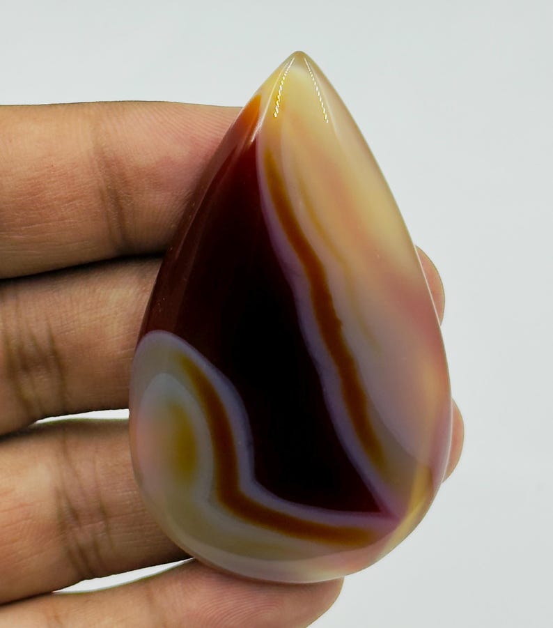 May include: A polished, teardrop-shaped agate gemstone featuring smooth, flowing bands of deep maroon, translucent cream, and soft amber. The stone has a glossy, reflective surface with distinct, organic color transitions throughout its structure.