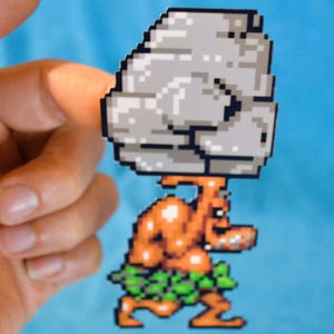 Peut inclure: Autocollant pixélisé représentant une figurine orange musclée portant un rocher gris sur la tête. La figurine a des feuilles vertes autour de la taille et est en mouvement. Le fond est bleu uni.
