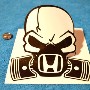 Può includere: Un adesivo a forma di teschio bianco e nero con il logo Honda al centro. Il design è minaccioso, con forme a pistone su entrambi i lati del logo. Sono visibili anche un piccolo oggetto dorato e un giocattolo a forma di dinosauro.