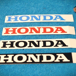 Peut inclure: Quatre autocollants rectangulaires Honda en bleu, rouge, noir et noir avec lettrage blanc. Les autocollants sont sur une surface bleue, avec deux billes de verre transparentes et un jouet en forme de dinosaure.