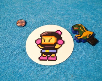 Könnte beinhalten: Ein pixeliger Charakteraufkleber auf einem weißen Kreis, ein kleiner Stein und ein USB-Stick in Dinosaurierform liegen auf einer blauen Oberfläche. Der Aufkleber zeigt einen Charakter mit rosa Boxhandschuhen und Stiefeln. Der USB-Stick ist grün und gelb.