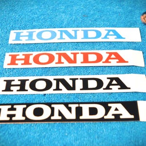 Op de afbeelding: Vier rechthoekige stickers met het woord HONDA in hoofdletters. De bovenste sticker heeft lichtblauwe tekst op een witte achtergrond, de tweede rode tekst op wit, de derde zwarte tekst op wit en de onderste witte tekst op een zwarte achtergrond.