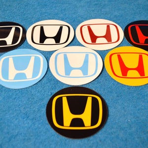 Puede incluir: Una colección de siete pegatinas circulares con el logotipo de Honda en varias combinaciones de colores, incluyendo blanco y negro, rojo y blanco, rojo y negro, azul claro y blanco, amarillo y rojo, y amarillo y negro, dispuestas en pirámide.