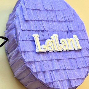 May include: A lavender piñata with the name "Leilani" in a light yellow font. The piñata is round with a ruffled texture and a black handle. The background is a light yellow.