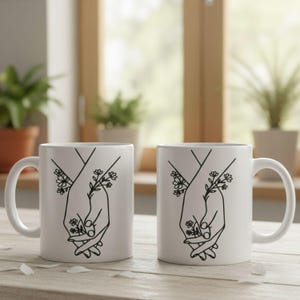 May include: Two white ceramic mugs with a black line drawing of two hands holding each other, with floral accents. The mugs have a curved handle and are set on a light-colored wooden surface.