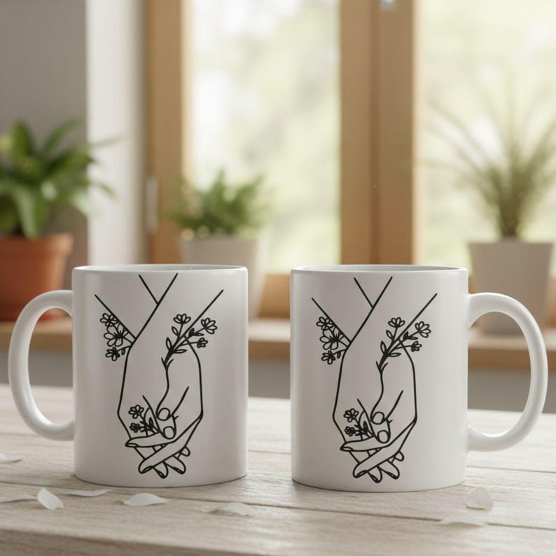 May include: Two white ceramic mugs with a black line drawing of two hands holding each other, with floral accents. The mugs have a curved handle and are set on a light-colored wooden surface.