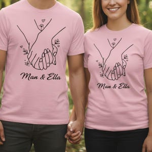 May include: Two light pink t-shirts with a black line drawing of two hands holding each other. The text "Man & Ella" is below the graphic. The shirts are short-sleeved.