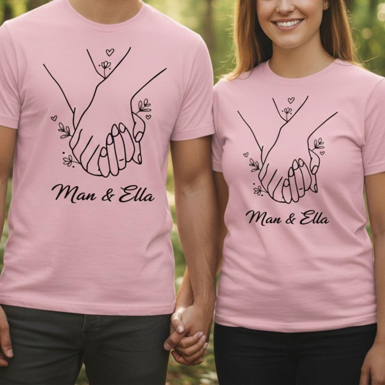 May include: Two light pink t-shirts with a black line drawing of two hands holding each other. The text "Man & Ella" is below the graphic. The shirts are short-sleeved.