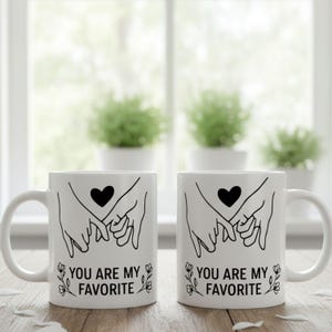 May include: Two white ceramic mugs with black line art. Each mug features a heart above clasped hands and the text "YOU ARE MY FAVORITE" with floral accents. The mugs are on a wooden surface.