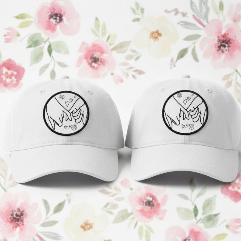 May include: Two white baseball caps with a curved brim. Each cap features a circular black and white patch with two hands holding each other's pinky fingers. The background is a floral pattern with pink and white flowers.