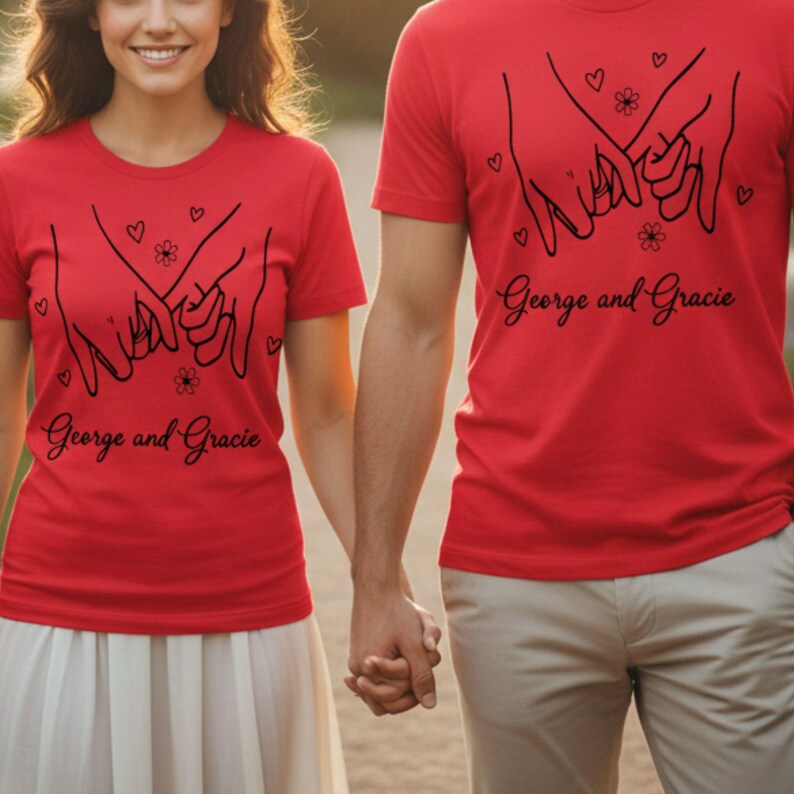 May include: Red t-shirts with a black line drawing of two hands holding pinky fingers, surrounded by hearts and flowers. The text "George and Gracie" is written in cursive below the hands. The shirts are worn by a couple.