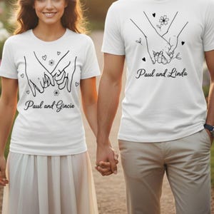 May include: White t-shirts with a black line drawing of two hands holding each other, with hearts and flowers. The text "Paul and Linda" and "Paul and Gracie" are printed on the shirts. The shirts are short-sleeved.