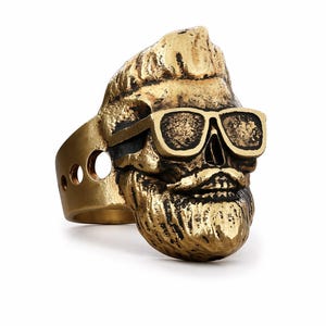 Puede incluir: Un anillo de color dorado que representa una calavera con gafas de sol y barba. El anillo tiene un dise&ntilde;o detallado, con las caracter&iacute;sticas de la calavera claramente visibles. La banda del anillo tiene tres recortes circulares. El anillo es probablemente de metal.