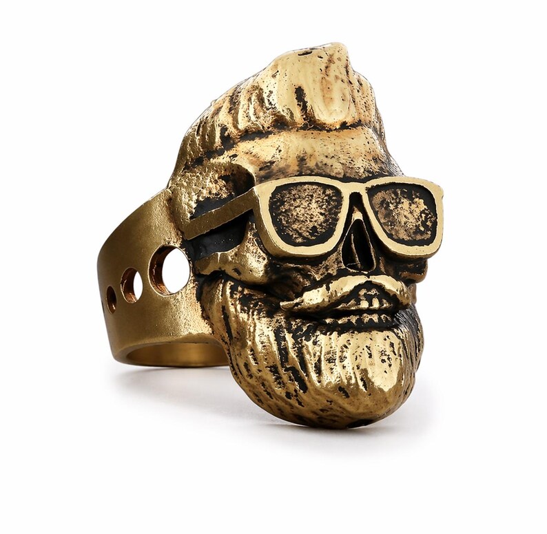 Puede incluir: Un anillo de color dorado que representa una calavera con gafas de sol y barba. El anillo tiene un dise&ntilde;o detallado, con las caracter&iacute;sticas de la calavera claramente visibles. La banda del anillo tiene tres recortes circulares. El anillo es probablemente de metal.
