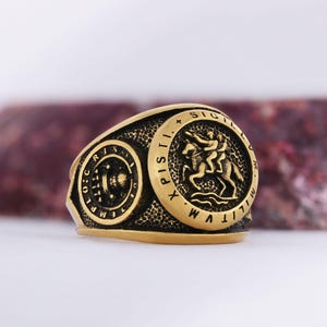 May include: A gold-colored ring with a detailed design. The ring features two circular emblems with inscriptions and a central emblem depicting two figures on horseback. The ring has a textured surface.