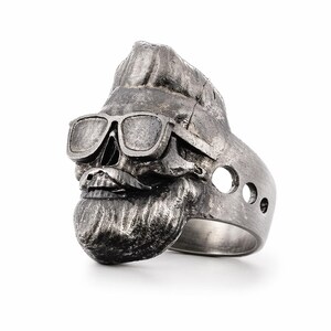 Puede incluir: Anillo de calavera plateado con gafas de sol, bigote y barba. El anillo tiene un acabado texturizado y tres peque&ntilde;os agujeros en el lateral. La calavera tiene un peinado tup&eacute;.