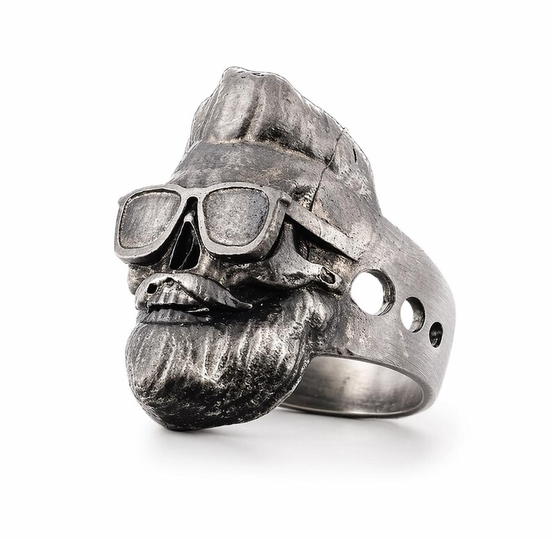 Puede incluir: Anillo de calavera plateado con gafas de sol, bigote y barba. El anillo tiene un acabado texturizado y tres peque&ntilde;os agujeros en el lateral. La calavera tiene un peinado tup&eacute;.