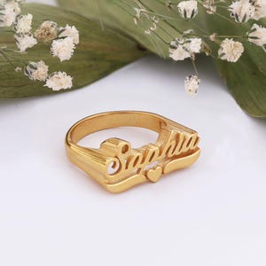 Puede incluir: Un anillo dorado con el nombre "Sophia" en cursiva. Un pequeño corazón está junto al nombre. El anillo está sobre una superficie blanca con vegetación y pequeñas flores blancas en el fondo.