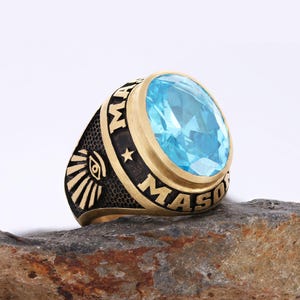 Puede incluir: Un anillo masónico dorado con una gran gema azul claro. El anillo presenta la palabra "MASON" en letras negras, una estrella y un símbolo de ojo. El anillo está sobre una superficie de piedra.