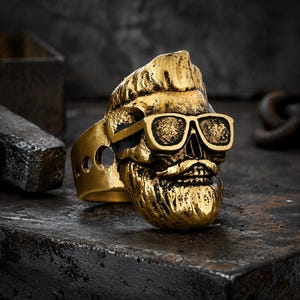 Puede incluir: Un anillo de calavera dorado con gafas de sol y barba. El anillo tiene un dise&ntilde;o detallado, con las caracter&iacute;sticas de la calavera claramente visibles. El anillo est&aacute; sobre un fondo oscuro y texturizado.