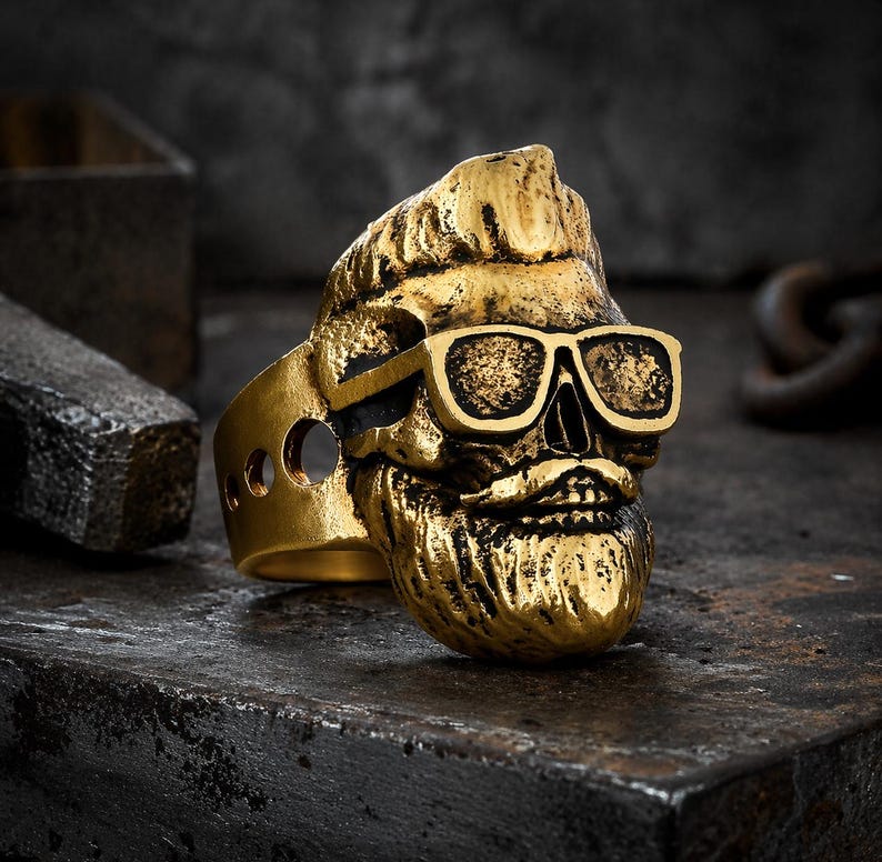 Puede incluir: Un anillo de calavera dorado con gafas de sol y barba. El anillo tiene un dise&ntilde;o detallado, con las caracter&iacute;sticas de la calavera claramente visibles. El anillo est&aacute; sobre un fondo oscuro y texturizado.