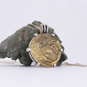 Könnte beinhalten: Goldfarbener Münzanhänger mit detailliertem Profil, eingefasst in einen silbernen Rahmen. Der Anhänger hängt an einer Silberkette. Die Münze wird vor einem rauen, grün-grauen Stein präsentiert.