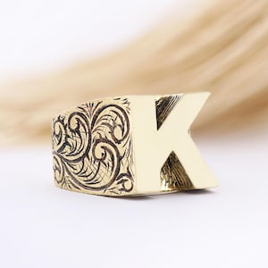 May include: A gold-coloured ring with the letter "K" as its main design. The ring has an engraved, swirling pattern on one side and a smooth, polished finish on the letter. The ring is set against a soft, neutral background.