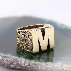 May include: A gold-coloured ring featuring the letter "M". The ring has an engraved floral pattern on the sides. The ring is set against a reflective surface.