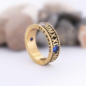 May include: A gold-colored ring with Roman numerals engraved around the band. The ring features three gemstones: two blue and one clear. The ring is a close-up shot, showcasing the details of the jewelry.