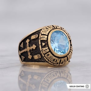 May include: A gold-toned ring featuring a large, round, faceted light blue gemstone at the center. The band is engraved with the Latin text AD ROSAM PER CRUCEM AD CRUCEM PER ROSAM and displays a cross symbol on the side with a dark, textured finish.