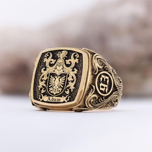 May include: A gold-colored signet ring with a detailed crest design. The ring features a rectangular face with a black background and the name "Adam" inscribed. Intricate scrollwork and a circular emblem adorn the sides, creating a classic, personalized accessory.