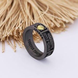 Puede incluir: Un anillo gris oscuro con la inscripción "JOSHUA" y "MASSACHUSETTS INSTITUTE OF TECHNOLOGY" grabada en la banda. El anillo presenta una piedra preciosa amarilla y las palabras "FOREVER TOGETHER".