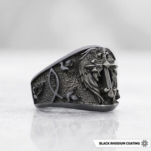 Puede incluir: Un anillo gris oscuro con revestimiento de rodio negro que presenta una cruz con alas en relieve, un símbolo de pez con las letras griegas IXOYE y manos en oración. La superficie metálica tiene un acabado mate y texturizado.