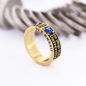 Może przedstawiać: Złoty pierścionek z niebieskim kamieniem szlachetnym. Pierścionek ma czarny napis "1897" i "San Diego State University". Widoczne są również rzymskie cyfry. Pierścionek jest prawdopodobnie pierścionkiem szkolnym lub pamiątkowym.