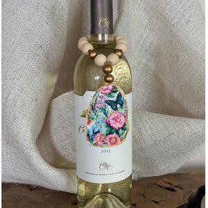 May include: A wine bottle with a floral label and a wooden bead wine charm. The label features pink roses, blue butterflies, and the year 2023. The wine charm has light wood beads and gold accents.