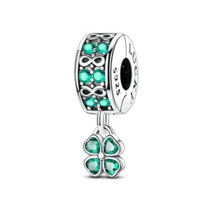 May include: A silver charm with a clover and infinity symbol design. The charm features a four-leaf clover pendant with green gemstones. The main body of the charm has a band with green gemstones and infinity symbols. The word "LUCKY" is engraved on the side.
