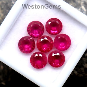 May include: A collection of seven round, faceted rubies in a white square container. The gemstones are a vibrant red color and are displayed against a dark, textured background. The text "WestonGems" is visible on the container.