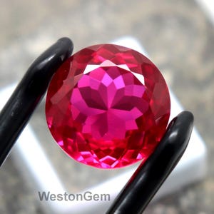 Può includere: Una gemma rosa brillante, rotonda e sfaccettata, è tenuta da pinzette nere. La gemma è posta su un quadrato bianco e una superficie grigia. Il testo "WestonGem" è visibile.